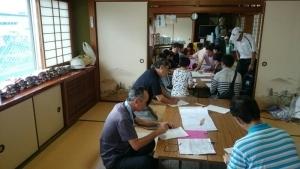  畳の部屋にテーブルが置かれていて、参加者達がテーブルの周りに座って資料を見ている写真