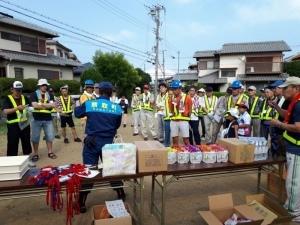 蛍光ベストをきた住民が、前方で説明をする作業着を着た男性の話を聞いている写真