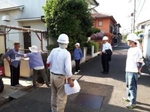 住宅街の道にヘルメット姿の参加者たちが立って訓練の様子を見ている写真