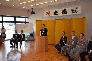 祝金婚式と壁に文字が貼られていて、金屏風の前に置かれた講演台で町長が話をしている写真