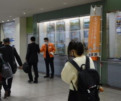 駅の通路でオレンジ色のジャンパーを着た町長が通行人にチラシを配っている写真