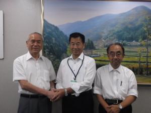 風景が描かれた絵画の前で握手をする向井通彦氏と町長、関係者の男性が並んでいる記念写真