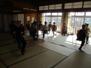 畳の部屋で参加者達が椅子に座って片足を上げて体操をしている写真