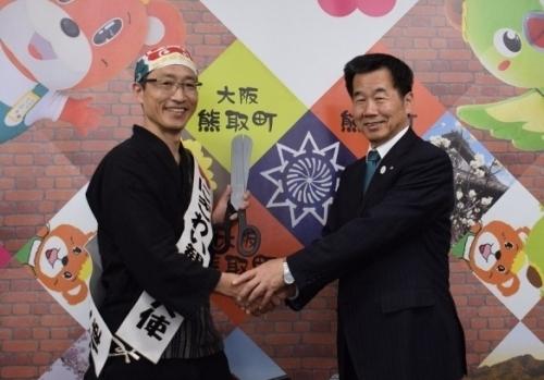 にぎわい観光大使のタスキをかけた山中慎介氏と町長が握手をしている写真