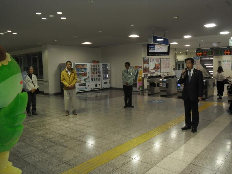 駅の改札口の前で町長がマイクを持って話をし、横に3人の男性が立っている写真