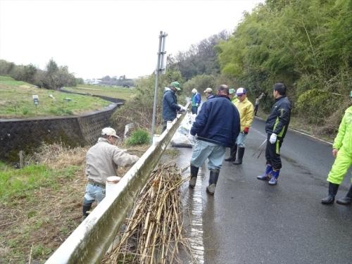 道路の脇にあるガードレールの周りでジャンパーを着た男性達が枝などを集めて清掃活動をしている写真