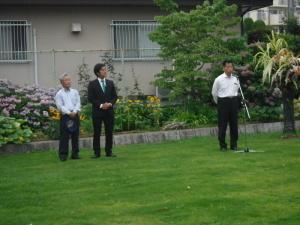 緑の芝の上で、マイクの前に立つ町長と、関係者の男性2名の写真