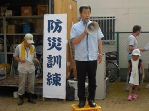 防災訓練と書かれた看板の前で、黄色い箱の上で、拡声器をもって話をする町長の写真