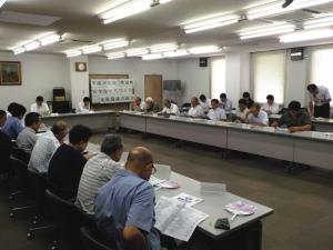 コの字の形に並んでいるテーブルに参加者達が座っていて、右側の男性がマイクを持って立って話をしている写真