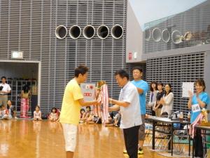 黄色のシャツを着た男性が町長からトロフィーを受け取っている閉会式の写真