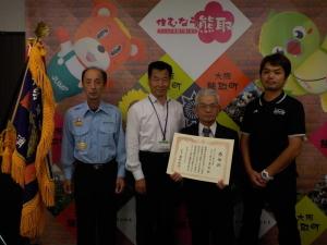 ジャンプ君とメジーナちゃんの描かれた壁の前で町長、庄司ゴルフ株式会社の関係者たちが並んでいる記念写真