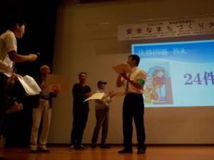 舞台の上で、書類を持ちながら講演をしている関係者の写真