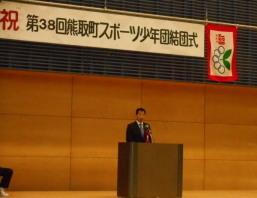 結団式で、リボン記章を付けた町長が演台に立っている写真
