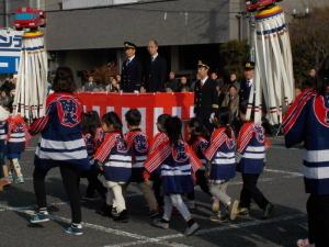 奥には紅白の幕がある台に乗った町長、手前には防災と書かれた法被を着た子供たちが歩いている写真