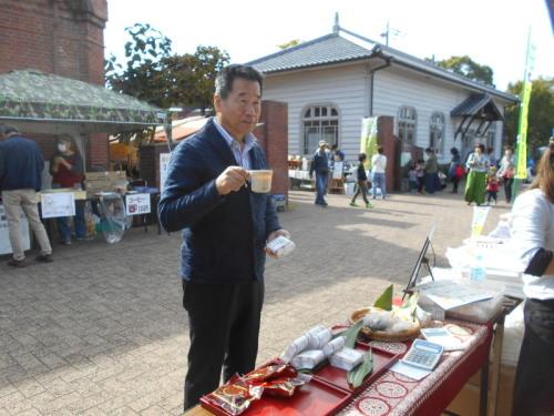 広場にコーヒーやお菓子などが売られた露店が出ていて、町長がマグカップを持ちながら立っている写真