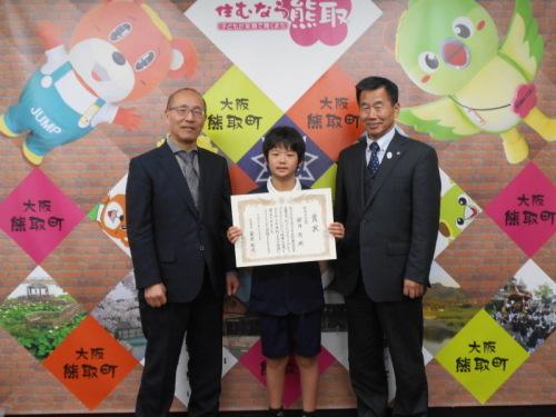 ジャンプ君とメジーナちゃんのが描かれた壁の前で、左から教育長、櫻井懸さん、町長が並んでいる記念写真