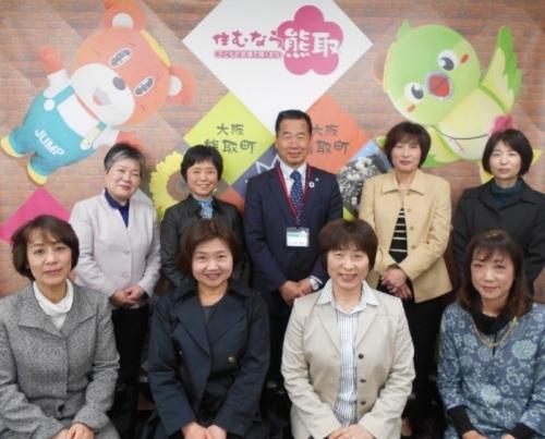 ジャンプ君とメジーナちゃんのが描かれた壁の前で、熊取町婦人会の女性8名と町長が並んでいる記念写真