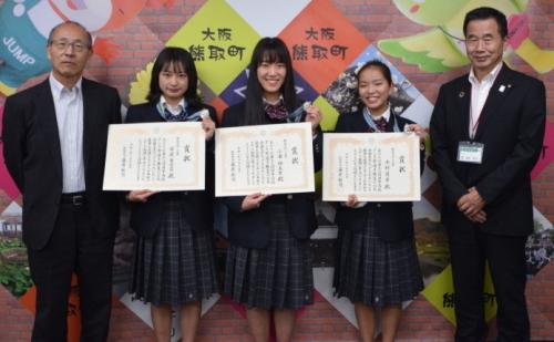 中央に首にメダルをかけ、賞状を持った制服姿の3人の女子生徒、右側に町長、左側に教育長が並んでいる記念写真