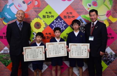 表彰状を持ち笑顔で立つ子供3人と、両脇に立つ町長と関係者男性の写真