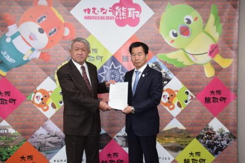 ジャンプ君とメジーナちゃんが描かれた壁の前で森口佳樹会長と町長が一緒に諮問書を持ち並んでいる写真