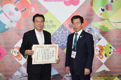 ジャンプ君とメジーナちゃんのが描かれた壁の前で感謝状を持った三原氏と町長が並んでいる写真