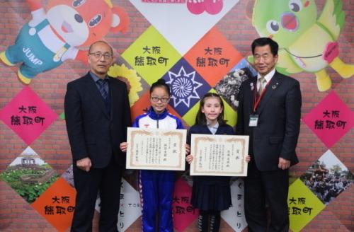 ジャンプ君とメジーナちゃんのが描かれた壁の前で教育長、表彰状を持った佐坂さんと佐藤さん、町長が並んでいる写真