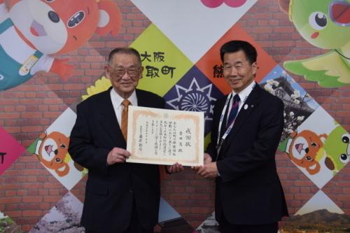 ジャンプ君とメジーナちゃんのが描かれた壁の前で感謝状を持った音田篤氏と町長が並んでいる写真