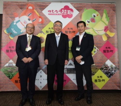ジャンプ君とメジーナちゃんが描かれた壁の前で町長と石本喜和男新会長、伊藤守副会長が並んで立っている記念写真