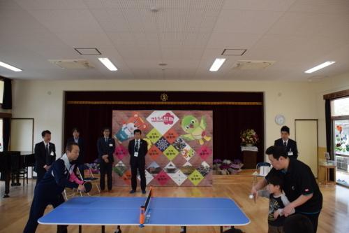 中央には卓球台があり向かい合って子供と大人が卓球をしていて、その様子を町長や関係者が見ている写真