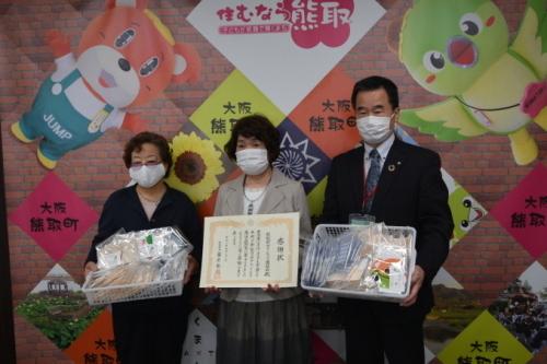 賞状を持った女性と、その両側で梱包された手作りマスク持つ町長と、女性の写真