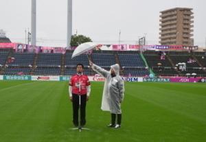 赤いユニフォームを着てマイクの前に立つ町長と、その横で町長に傘をさす関係者の写真