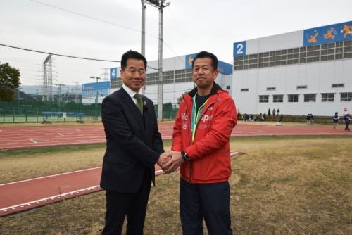 芝生のグラウンドの上で町長と山田村長が両手を重ねて握手をしている写真