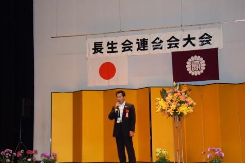 長生会連合会大会と書かれた幕が上にかかっていて金屏風が後ろに置かれている舞台で町長がマイクを持って大きく口を開けている写真