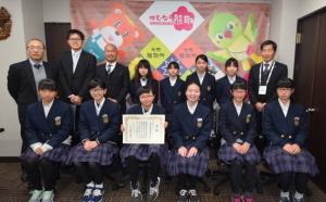ジャンプ君とメジーナちゃんのが描かれた幕の前で表彰状を持った女子学生たちと関係者、町長が並んでいる記念写真