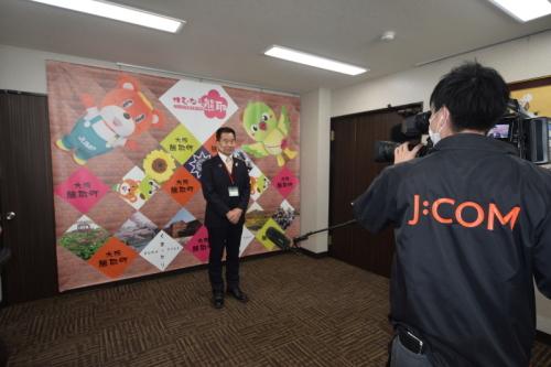 J:COMと書かれたジャンパーを着た男性が大きなビデオカメラで町長を撮影している写真