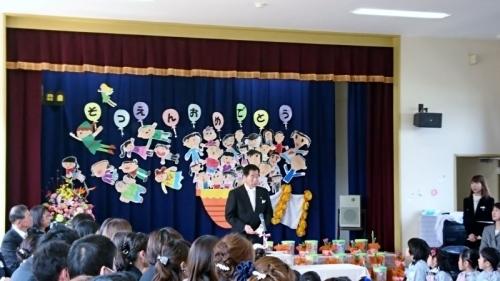 舞台上の幕には卒園おめでとうという文字と人の絵が貼ってあり、舞台の前で町長が子供たちに話をしている写真