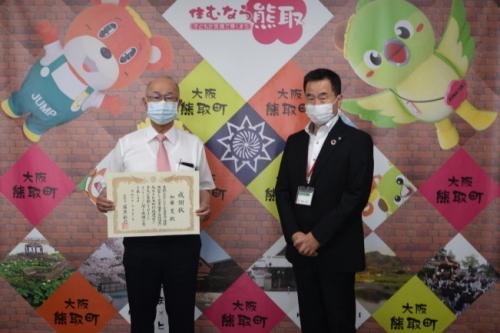 黒いスーツをきた町長とピンクのネクタイを着け賞状を持った男性が横に並んで立っている写真
