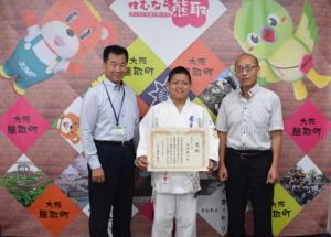 柔道着を着た市谷櫂人さんと、その左右に立つ町長と関係者の男性の写真