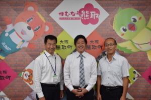 ジャンプ君とメジーナちゃんの描かれた壁の前で左から町長、宮原翔平さん、教育長が並んでいる記念写真