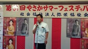 演歌歌手のポスターが貼られた紅白の幕の前で町長がマイクを持って話をしている写真