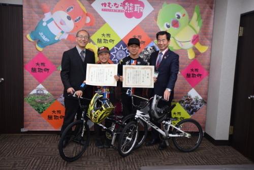 ジャンプ君とメジーナちゃんのが描かれた壁の前で町長と教育長、表彰状を持った北川晃久君、北川晋也君、自転車2台が写っている写真