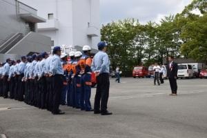 アスファルトの敷地に消防団員たちが横二列に並んでいて、団員たちの前でスーツ姿の町長が話をしている写真