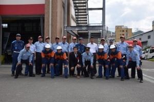 アスファルトの敷地に制服や作業着を着た消防団員たちと町長が横2列に並んでいる記念写真