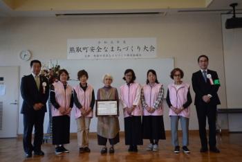 中央に賞状を持った女性、両脇にピンクのベストを着た女性達や町長が並んでいる写真