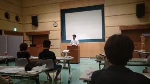 大きなスクリーンの前に置かれた講演台で、町長が座っている参加者達に向かって話をしている写真