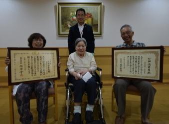 車いすに座った方の後ろに町長が立ち、その両側に座った方々が額縁をこちらに向けて撮影されている写真