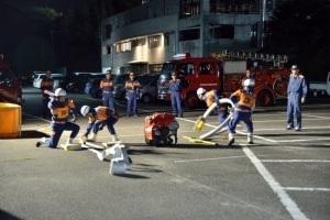  奥には消防車が停まっていて、手前では消防団員たちがポンプのホースを持ち訓練をしている写真