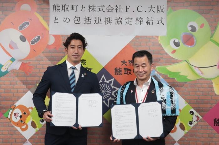 肩にタオルをかけた町長とスーツ姿の男性がお互いに協定書を持って立っている写真