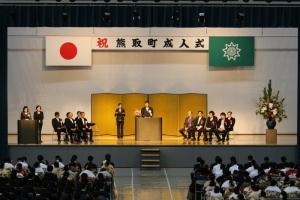 祝熊取町成人式と書かれている横断幕の下で、後ろに金屛風のある壇上で町長が話をしていて、晴れ着姿の参加者達が話を聞いている写真