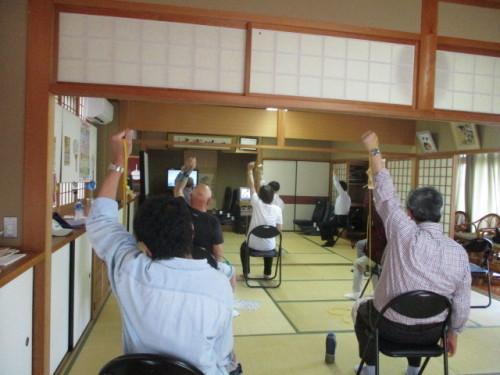 畳の部屋の中で参加者達が椅子に座って左手を上げて体操をしている様子を後ろから写している写真
